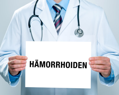 Arzt hält weißen Zettel in der Hand auf dem das Wort "Hämorrhoiden" steht.